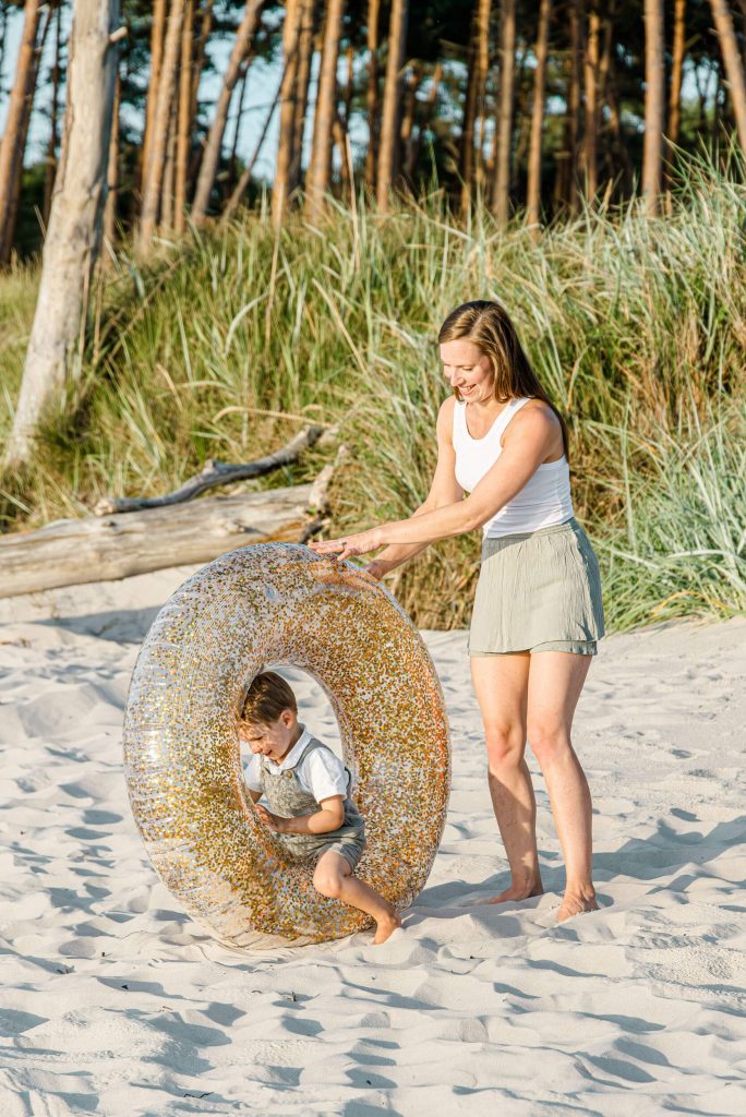 Eine Mutter hilft ihrem Kind mit einem Schwimmreifen am Strand, umgeben von Sand und Vegetation. Familienzeit am Meer.