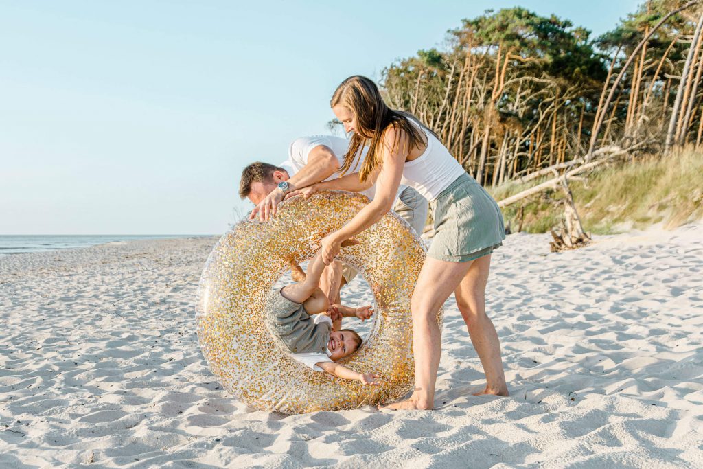 Familie mit einem Kind in einem glitzernden Schwimmreifen am Strand, umgeben von Sand und Bäumen, bei Sonnenlicht.