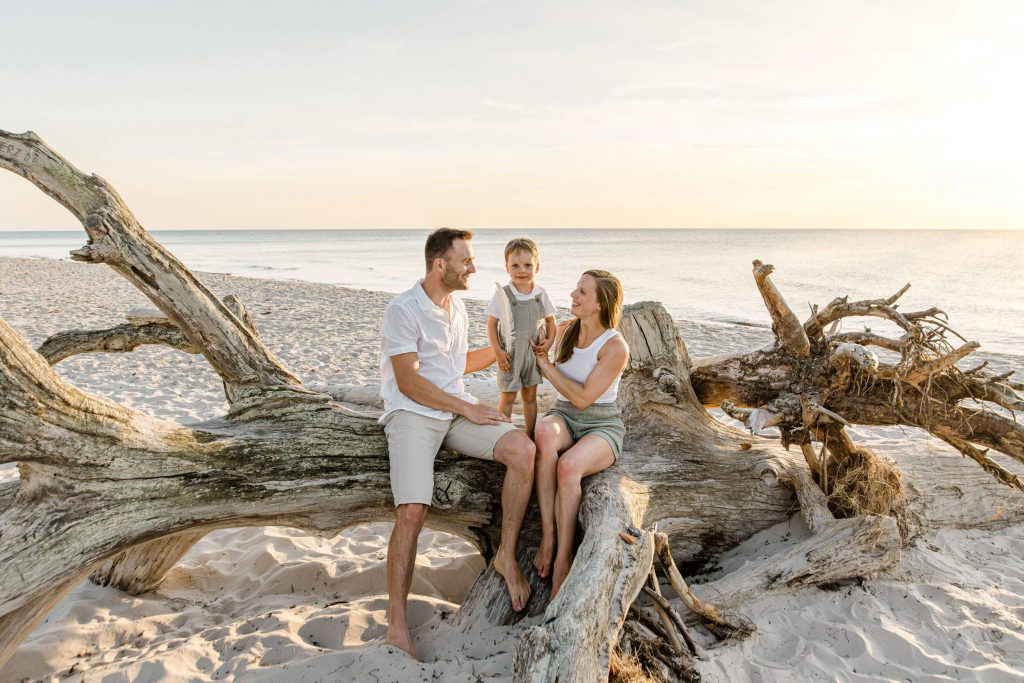 Familie sitzt auf einem Baumstamm am Strand, lächelnd und entspannt, mit einem Kind in der Mitte, umgeben von Sand und Wasser.