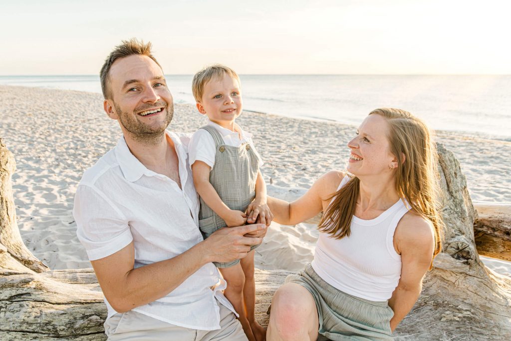 Familienfoto am Strand mit einem lachenden Paar und einem kleinen Jungen, der zwischen ihnen sitzt, vor einer ruhigen Meereslandschaft.