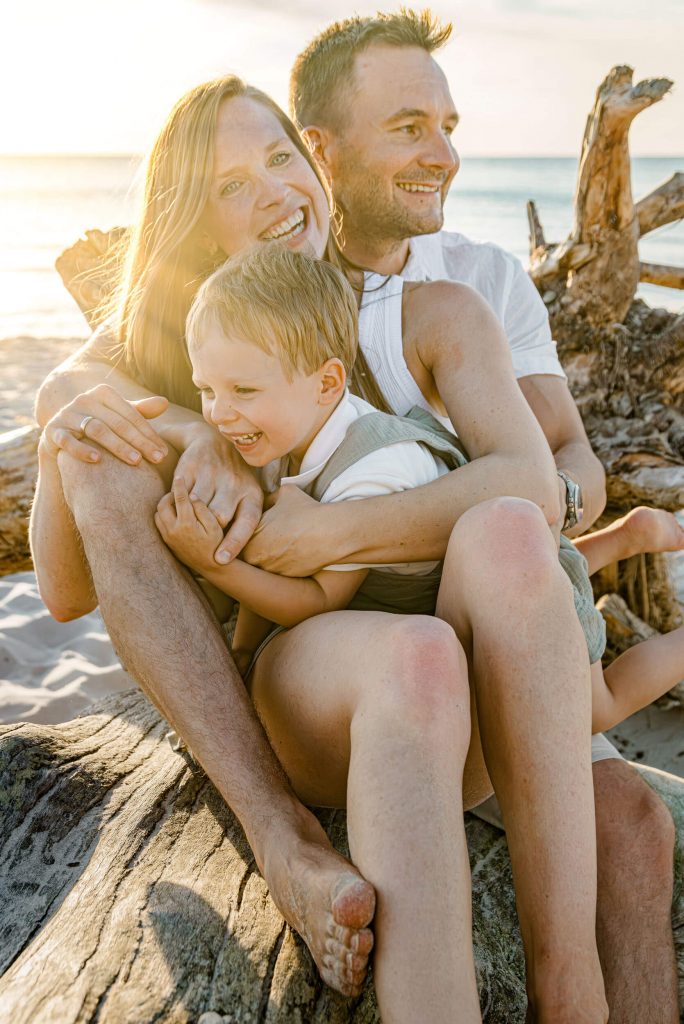 Familie sitzt auf einem Baumstamm am Strand, lachend und umarmend, mit Sonnenlicht im Hintergrund, ideal für Familienfotos.