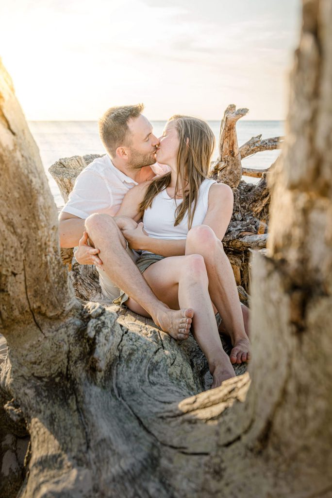 Ein Paar küsst sich am Strand, umgeben von Holzstämmen, während die Sonne untergeht. Romantische Atmosphäre für Familienfotos.