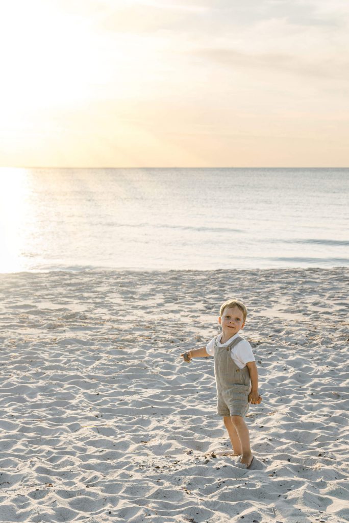 Ein kleines Kind in einem hellen Outfit steht am Strand und blickt zur Seite, während die Sonne untergeht. Sand und Wasser im Hintergrund.