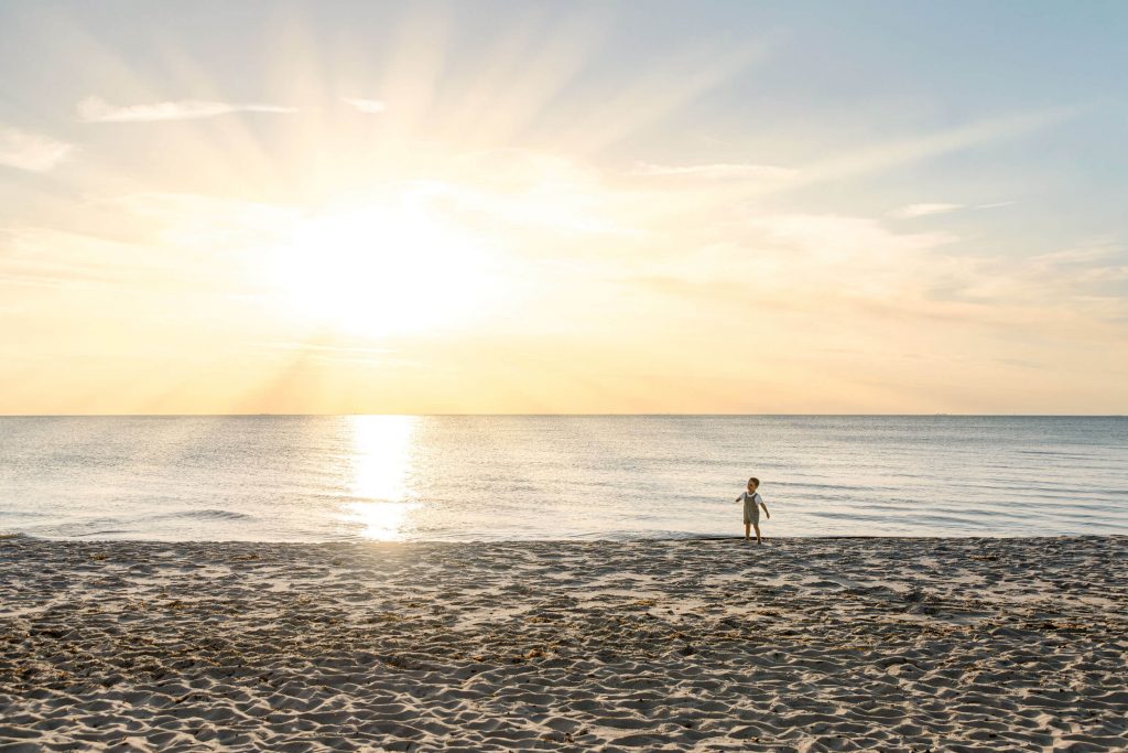 Ein Kind steht am Strand, während die Sonne über dem Wasser untergeht. Der Himmel ist klar mit sanften Farben.