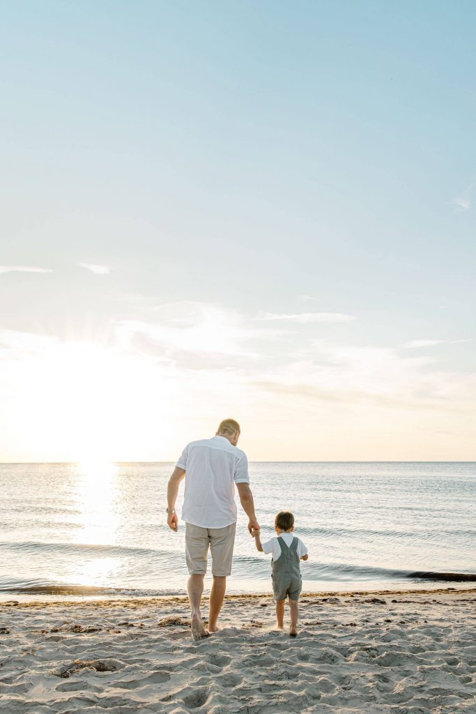 Ein Vater und sein Kind gehen am Strand entlang, während die Sonne untergeht. Familienzeit am Meer mit ruhiger Wasseroberfläche.