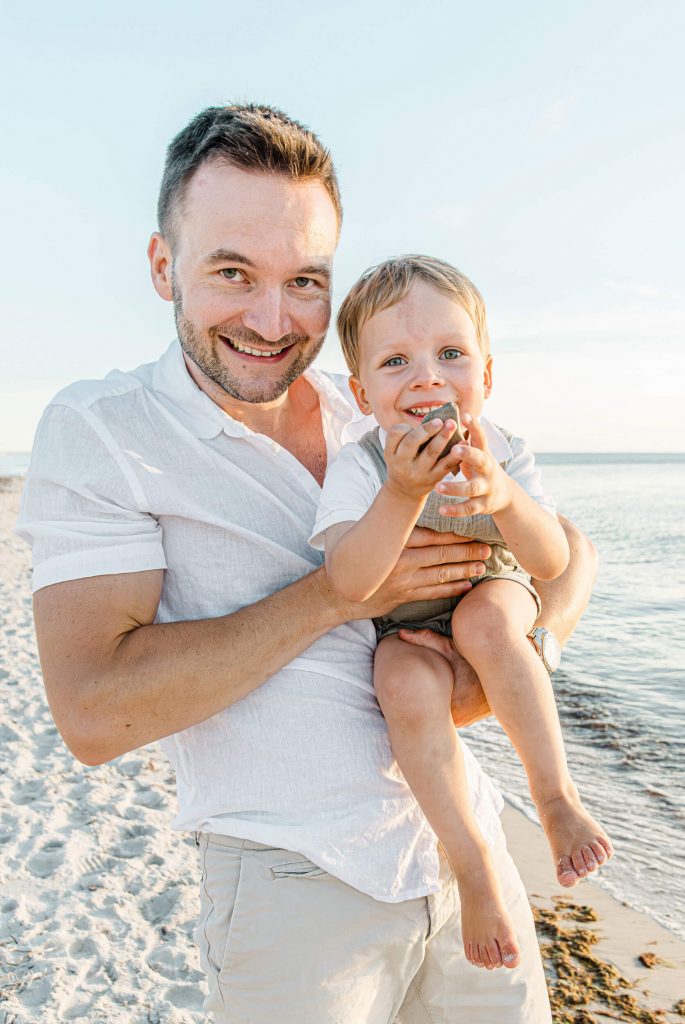 Ein Vater hält seinen kleinen Sohn am Strand, beide lächeln glücklich. Familienfoto am Meer mit Sand und Wasser im Hintergrund.