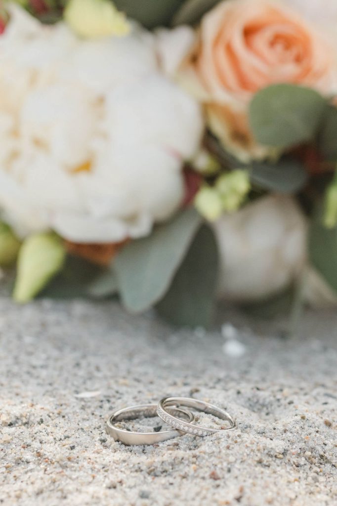 Zwei Eheringe auf Sand vor Blumenstrauß