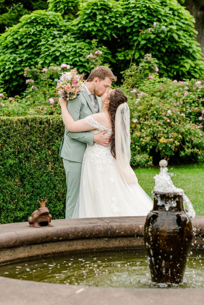 Brautpaar umarmt sich im Garten am Springbrunnen