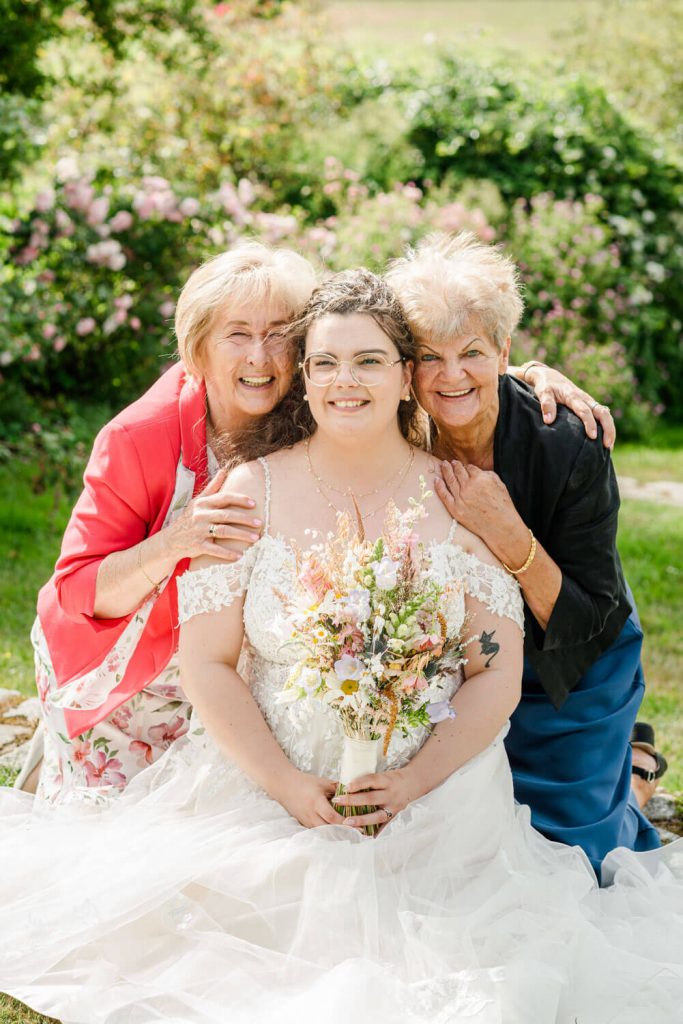 Braut mit Blumenstrauß und älteren Damen draußen.