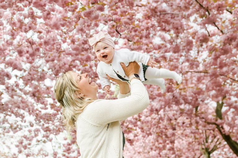 Mutter hebt Baby unter Kirschblütenhimmel lachend hoch